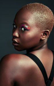 Femme avec une coupe très courte aux cheveux châtain cuivré, regard intense par-dessus l’épaule, maquillage artistique coloré autour des yeux.